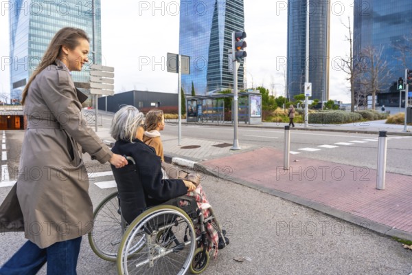 Young woman pushing elderly grandmother in a wheelchair while a child walks beside them across a