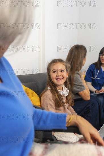 Smiling young girl visits grandmother in a wheelchair at a nursing home, surrounded by family and a