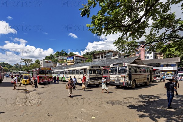 Busy bus stop with people and buses under a sunny sky with green trees, Kandy bus station in Sri