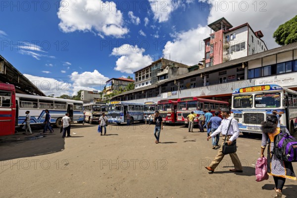 People walk past a bus stop surrounded by various buses and clouds in the sky, Kandy bus station in