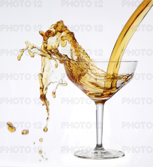 A strong flood of golden yellow liquid pours into a cocktail glass and spills over, studio shot