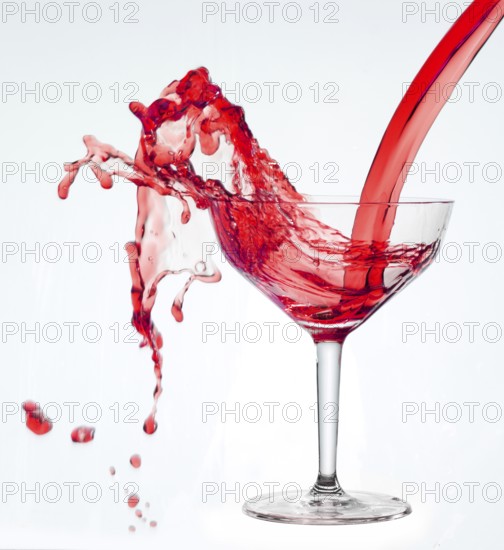 A powerful flood of red liquid pours into a cocktail glass and spills over, studio shot against a