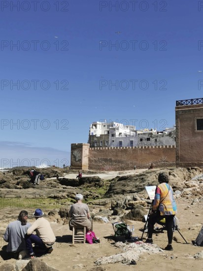 Painters paint the city view on the coast in front of old city walls and rocks under a blue sky,