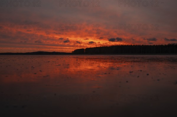 A dramatic sunset over a still, frozen lake and forest with orange-red hues in a cloudy sky,