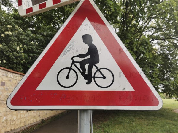 Road sign for bicycles, red and white triangle in the foreground, trees in the background, Czech