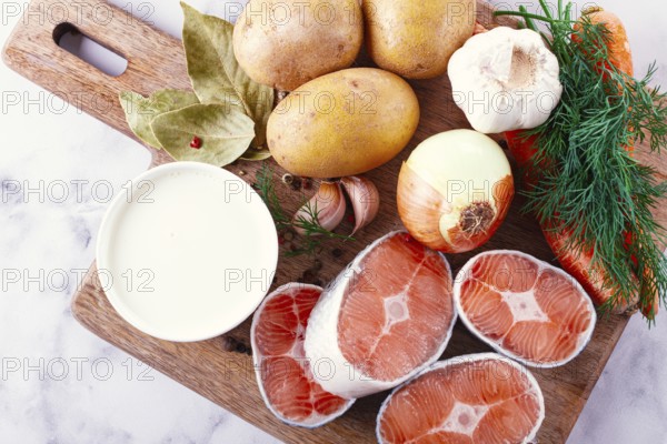 Potatoes, salmon, garlic, onion, and herbs are placed on a wooden cutting board. These ingredients