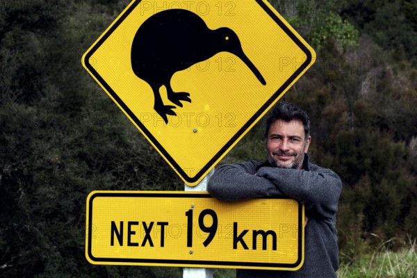 Warning sign with kiwi symbol on a road with a man in the background, Franz Josef Glacier, New