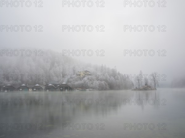 View across Königssee to boathouses, Christlieger island and frozen trees in fog, Schönau am