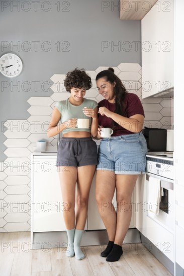 A happy lesbian couple stands in a kitchen, chatting and sharing a morning coffee. They enjoy