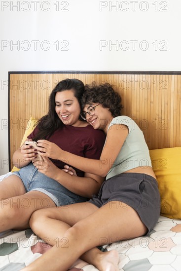 Two lesbian women sitting on a bed, enjoying a relaxed moment with a smartphone and a notebook.