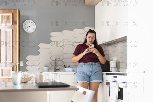 A curvy woman in a casual outfit stands in a modern kitchen, intently using her smartphone. The