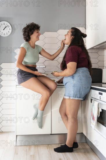 A happy lesbian couple shares a playful moment in a modern kitchen. One partner sits on the