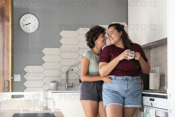 A happy lesbian couple shares a playful moment in a modern kitchen. One partner sits on the