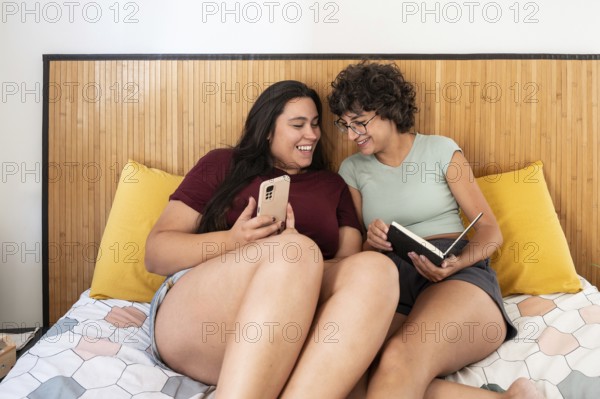 Two lesbian women sitting on a bed, enjoying a relaxed moment with a smartphone and a notebook.