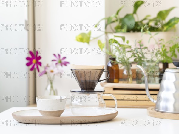 Modern pour over coffee brewing setup sits on a white table, with houseplants and flowers adding a