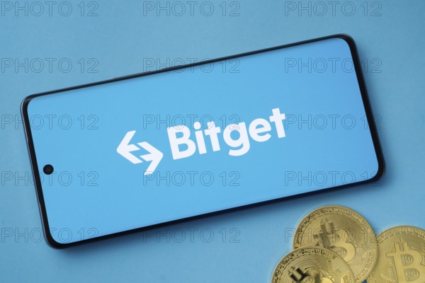 Dhaka, Bangladesh- 27 July 2025: Bitget logo displayed on a smartphone with Bitcoin coins on a blue