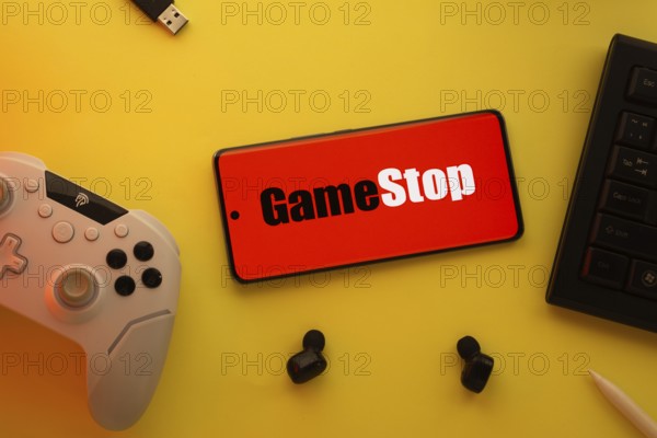 Dhaka, Bangladesh- 15 July 2025: Smartphone with GameStop logo, game controller, and earbuds on a