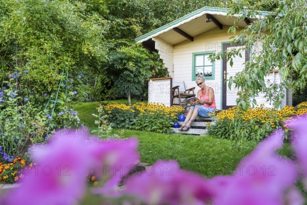 A woman sits relaxed on the small veranda of a garden shed and reads. Blooming summer flowers in