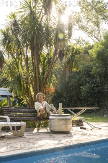 Elderly woman enjoys a peaceful moment in her backyard, sitting on a bench next to a sparkling