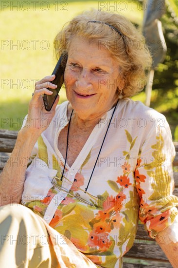 Elderly woman with glasses hanging from a string around her neck is sitting on a wooden bench