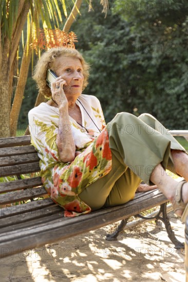 Senior woman sitting comfortably on a park bench, enjoying a sunny day while making a phone call.