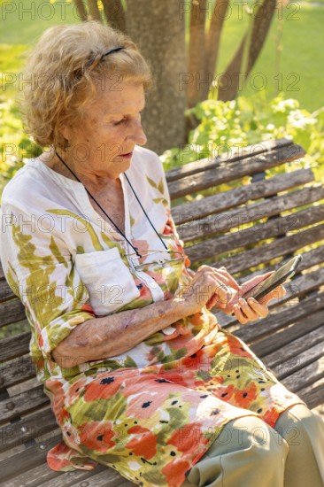 Senior woman sitting on a wooden bench in a park, enjoying the warmth of summer while using her