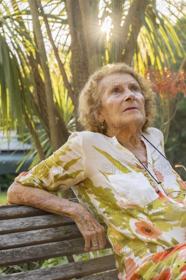 Senior woman enjoying a tranquil moment in a lush garden, sitting on a wooden bench while sunlight