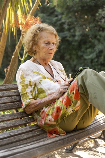 Senior woman sitting comfortably on a wooden bench in a park, using a tablet while enjoying the