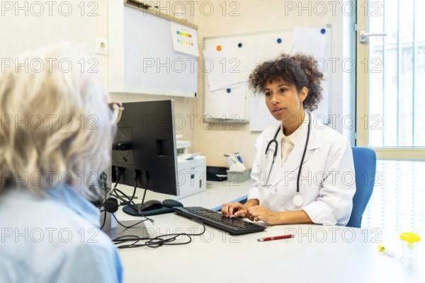 Female doctor using computer and listening to senior patient describing symptoms in hospital room