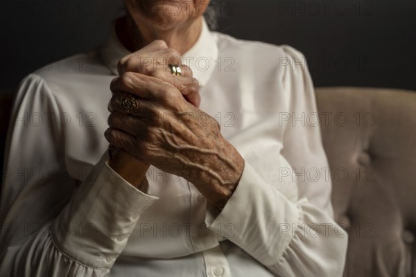 90 year old woman in white blouse holding folded hands, conveying thoughtfulness in a dim light,
