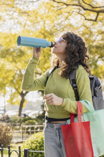 Woman with curly hair and sunglasses drinks from a blue water bottle in a park. She carries