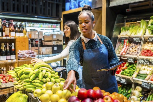 Supermarket employee using digital tablet while arranging fresh fruit on display, customer choosing