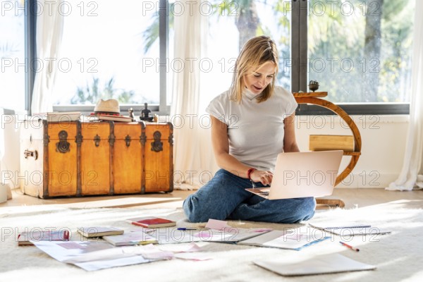 A woman sits on a bright home floor, absorbed in her laptop, with papers scattered around her.