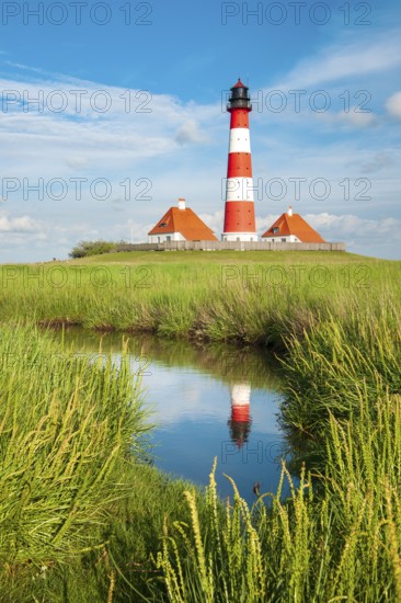 The Westerheversand lighthouse under a blue sky with white clouds reflected in a tideway,
