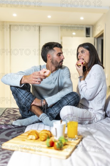 Young couple wearing pajamas sitting in bed eating donuts for breakfast, with croissants, orange