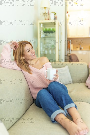 Vertical photo of a blonde mature adult happy relaxed woman drinking tea lying on the sofa