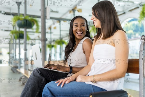 Two young businesswomen are sitting on a bench, working on a laptop and talking, in a light and