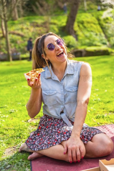 Cheerful young woman laughing while eating pizza in a vibrant green park, enjoying a sunny spring