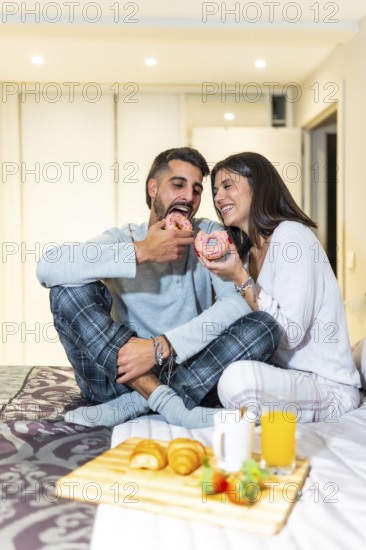 Young couple wearing pajamas, sitting in bed and eating breakfast, including donuts, croissants,