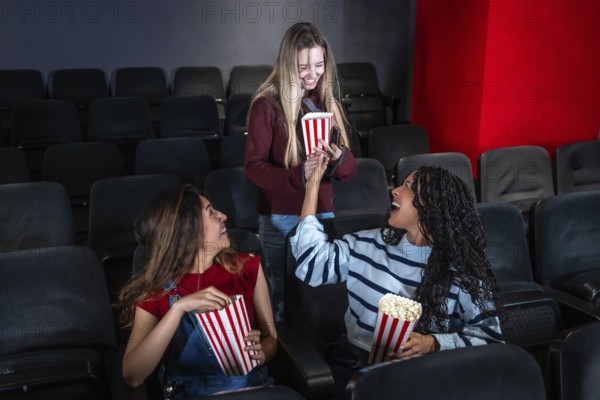 Three female friends are laughing and sharing popcorn while watching a movie in a cinema, enjoying