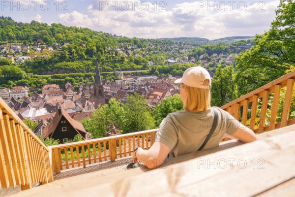 A person sitting on a terrace with a view of a town and the surrounding hills, Panorama Holzsteg,