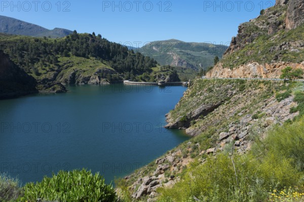 A tranquil lake in a mountainous landscape with green hills and rocks, under a clear blue sky,