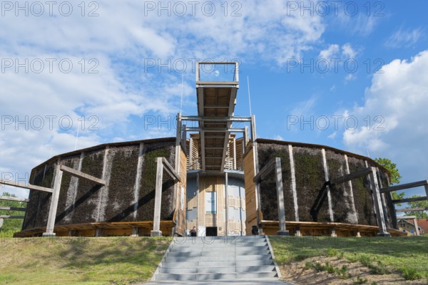 Round building made of wood and metal with stairs, blue sky and green landscape in the background,