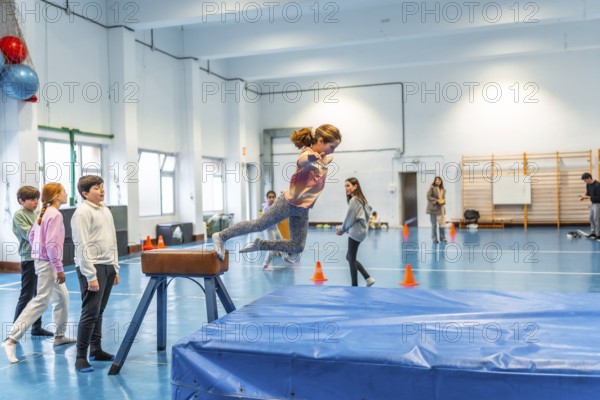 Elementary school girl practicing gymnastics, jumping over a pommel horse in the school gym watched