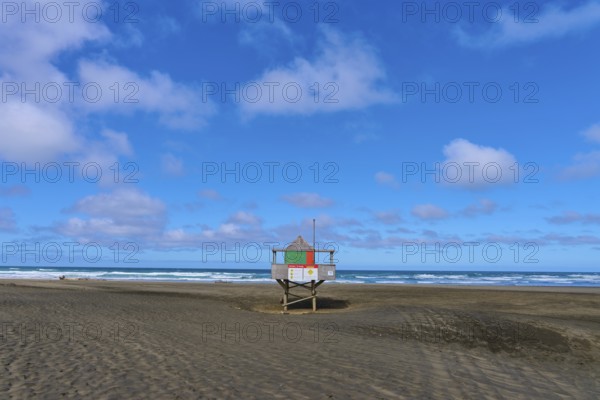 A rescue tower stands alone on a wide sandy beach under a blue sky, Bethells Beach, Te Henga,