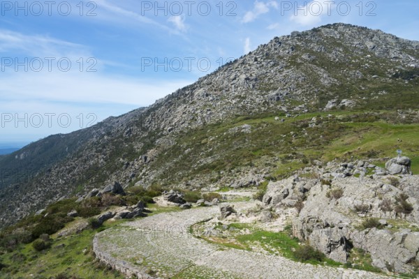 A winding road leads through a rocky landscape with green vegetation and high mountains in the