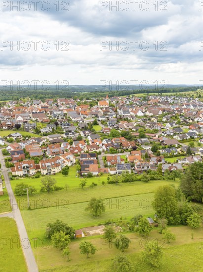 Aerial view of a village with red roofs, surrounded by green meadows and trees under a cloudy sky,