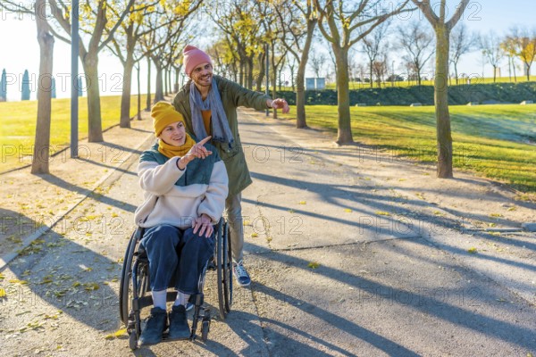 Two friends are walking in a park, one is using a wheelchair and they are both pointing at