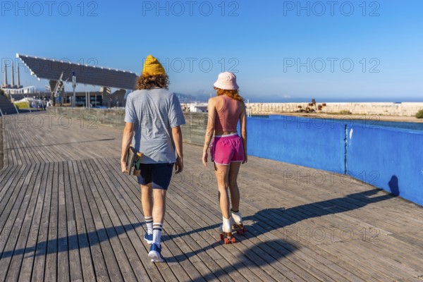 Young man carrying a skateboard strolls alongside a roller skating woman on a wooden pier, enjoying