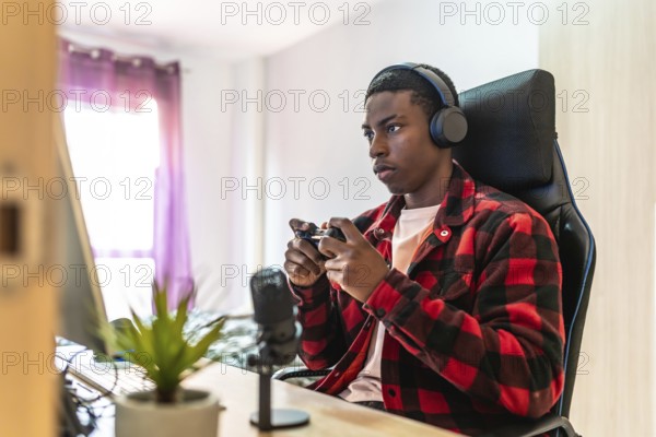 Young man playing videogames on a desktop computer using a controller, wearing headphones and using
